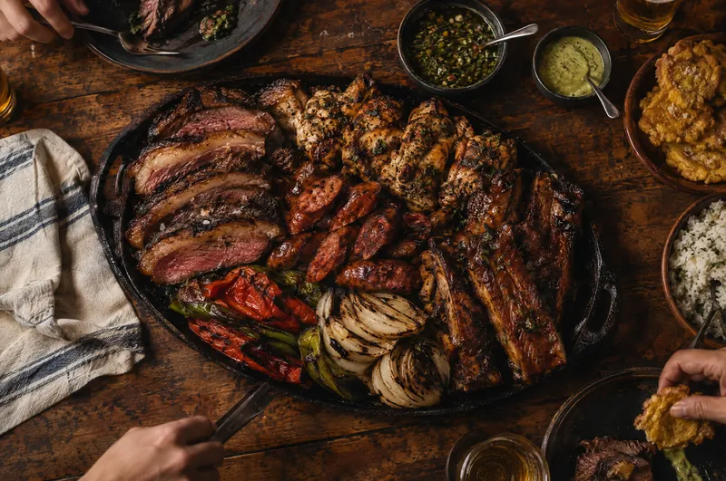 Overhead view of the Parrillada Familiar: sliced picanha, chicken, chorizo, ribs and grilled vegetables on an iron platter, hands reaching in
