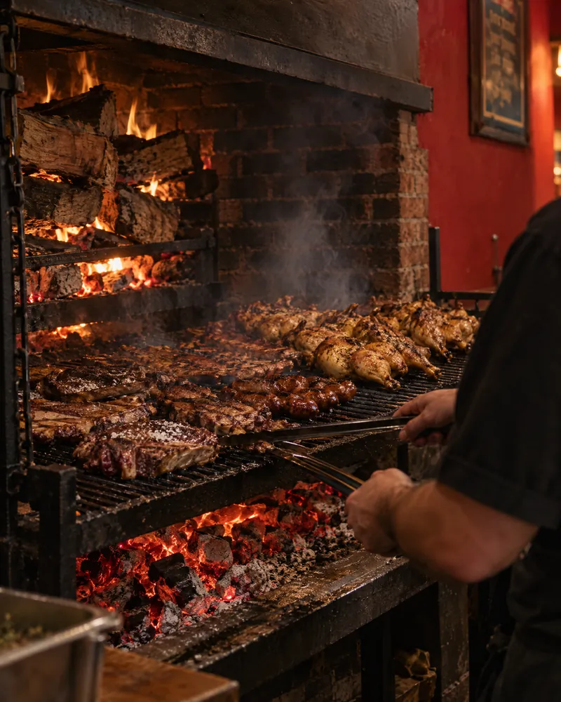 A cook’s hands turning steaks with long tongs on the iron grill, whole chickens and chorizo roasting over glowing oak coals